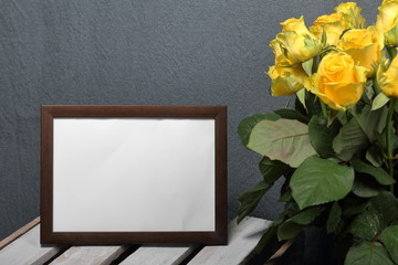 A large bouquet of yellow roses in a glass vase. Nearby is a wooden box. On the box is a beautiful frame for a photo.