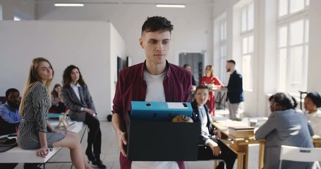 Sad young male handsome European manager is fired and quitting job at multiethnic office, walking away with stuff box.