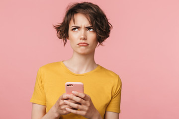 Confused young beautiful woman posing isolated over pink wall background using mobile phone.