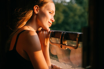 Mood atmospheric lifestyle portrait of young beautiful blonde hair girl looking out of window from riding train. Pretty teen enjoying beauty of nature from moving train car in summer. Travel concept