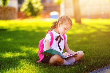 Little school girl with pink backpack sitting on grass after lessons and read book or study lessons, thinking ideas, education and learning concept.