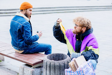 Squeamish handsome beard man sits on bench with smartphone while strange homeless man in colorful clothes rummaging in trash container in park gray background.