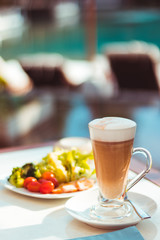 Tall glass of latter coffee on a table in ourdoor area next to swimming pool, hot drink and breakfast including salad