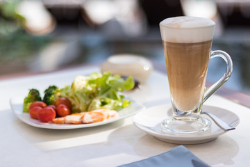 Tall glass of latter coffee on a table in ourdoor area next to swimming pool, hot drink and breakfast including salad