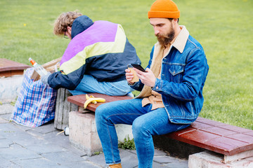 Hansome beard man with shock emotion sitting on the bench with disposable cup and smart phone while strange homeless man in colorful clothes rummaging in trash container in park green background.
