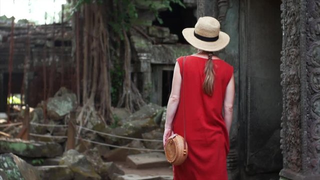 Slow motion: caucasian woman in red dress is walking among ruins of beautifully decorated Ta Prohm temple and taking photos. Angkor Wat complex. Cambodia