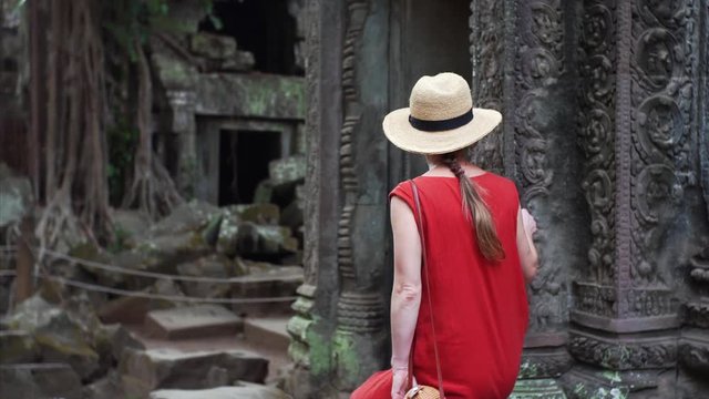 Following caucasian woman in red dress walking among ruins of beautifully decorated Ta Prohm temple in Angkor Wat complex. Cambodia
