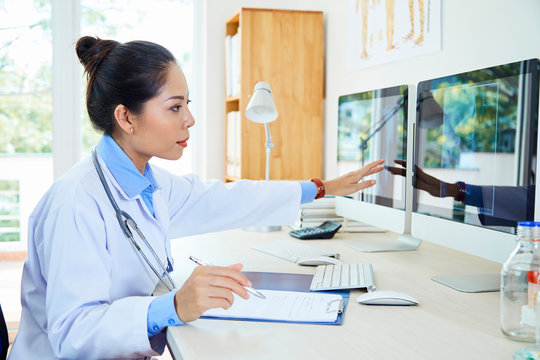 Asian young female radiologist sitting at the table working with documents and pointing at computer monitor with x-ray images at office