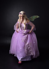 full length portrait of a blonde girl wearing a fantasy fairy inspired costume,  long purple ball gown with fairy wings,   standing pose on a dark studio background.