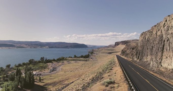 A drone flyby of the Columbia River landscape, from the mountainous road, towards the wide river, with a view of the mountain range behind in the horizon.
