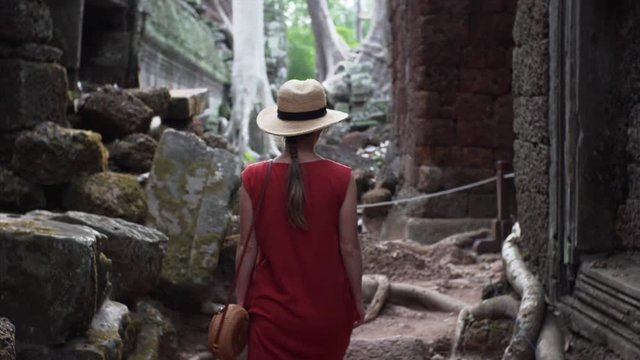 Following a caucasian woman in red dress walking among ruins of beautifully decorated Ta Prohm temple. Angkor Wat complex. Cambodia