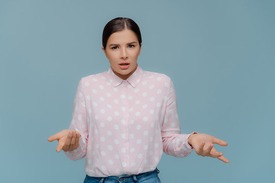 Pretty dark haired woman with makeup, spreads hands and looks doubtfully, feels misunderstanding, wears polka dot shirt and jeans, cannot make decision, poses indoor over blue studio background