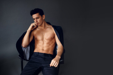 Fototapeta premium Elegant shirtless brunette man with jacket on his shoulders, seated on chair, on grey background.