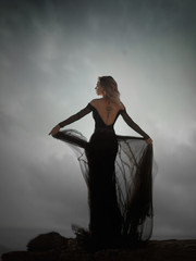 Fototapeta premium Young woman in long transparent black dress among beach and sea. Wind blows hem of her dress.