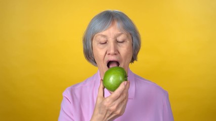 Portrait of cheerful senior woman with grey hair standing isolated on yellow background and eating green apple while smiling and looking at camera