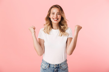 Happy young blonde woman posing isolated over pink wall background make winner gesture.