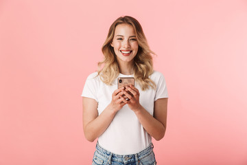 Happy young blonde woman posing isolated over pink wall background using mobile phone.