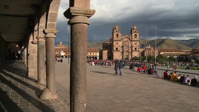 Wide low-angle still shot of Cathedral's cloister pillars, and surrounding street church buildings and Plaza de Armas with visitors relaxing at the park, Cusco, Peru