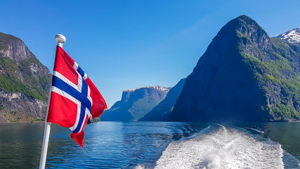 Norwegian flag hanging on  the railing of the ship and waving above the water.The motor of the ship makes the water wavy and foamy. Tall, lush green mountains surrounding the fjord. Clear blue sky. - obrazy, fototapety, plakaty
