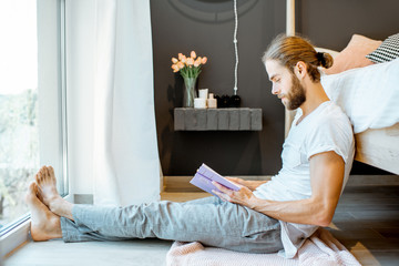 Handsome man in pajamas reading book while relaxing on the floor near the bed at home