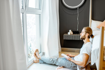 Handsome man in pajamas reading book while relaxing on the floor near the bed at home
