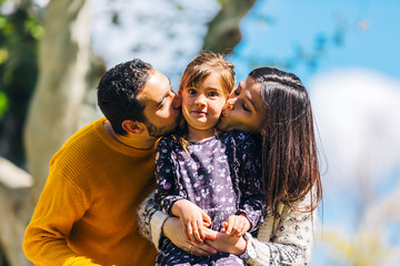 Fototapeta premium Front view of happy parents kissing her lovely daughter outdoors in the park in a sunny day