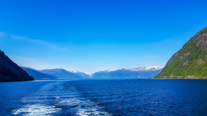 A view on the Songefjorden fjord from the water level. The motor of the ship makes the water wavy and foamy. Tall, lush green mountains surrounding the fjord. Clear blue sky.