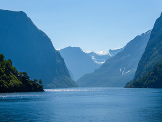 A view on the Songefjorden (King of the Fjords) from the water level. It is the deepest fjord in Norway. Tall, lush green mountains surrounding the fjord. Calm surface of the water. Clear blue sky.