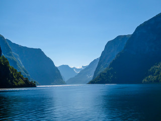 A view on the Songefjorden (King of the Fjords) from the water level. It is the deepest fjord in Norway. Tall, lush green mountains surrounding the fjord. Calm surface of the water. Clear blue sky.