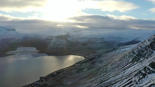 Nordic winter from helicopter view: Drone descends down side of impressive mountain range revealing sea inlet / lake reflecting sunset and sun flare in Isafjordur, westfjords, Iceland.