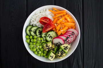 Vegan poke bowl with white rice and vegetables in the white  bowl in the center of the black wooden background.Top view.Closeup.