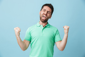 Happy excited young handsome bearded man posing isolated over blue wall background make winner gesture.