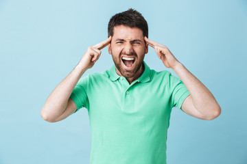 Stressed young handsome bearded man posing isolated over blue wall background screaming because of headache.