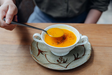 Hand scooping Classic Pureed Pumpkin Soup topping with pepper. Served in ceramic cup on wooden table.