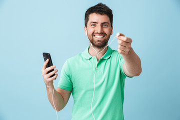 Happy young handsome bearded man posing isolated over blue wall background using mobile phone listening music with earphones.