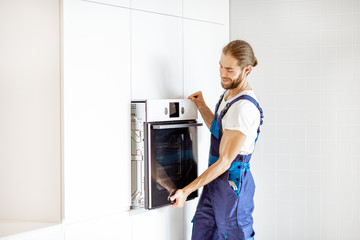 Handsome workman in workwear installing electric oven into the kitchen shelves at the modern kitchen at home