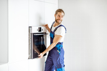 Fototapeta na wymiar Handsome workman in workwear installing electric oven into the kitchen shelves at the modern kitchen at home