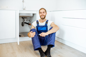 Portrait of a handsome plumber in overalls sitting near the sink at the kitchen