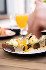 Morning breakfast with friends in a cozy summer outdoor cafe. Poached eggs on toasted black bread with avocado and Dutch sauce. Hands in the frame with golden cutlery.