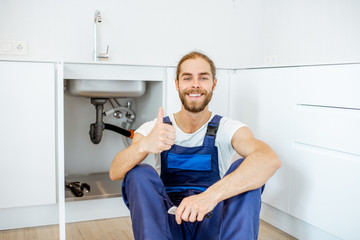 Portrait of a handsome plumber in overalls sitting near the sink at the kitchen