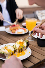 Morning breakfast with friends in a cozy summer outdoor cafe. Poached eggs on toasted black bread with avocado and Dutch sauce. Hands in the frame with golden cutlery.