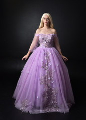 Fototapeta na wymiar full length portrait of a blonde girl wearing a fantasy fairy inspired costume, long purple ball gown with fairy wings, standing pose on a dark studio background.