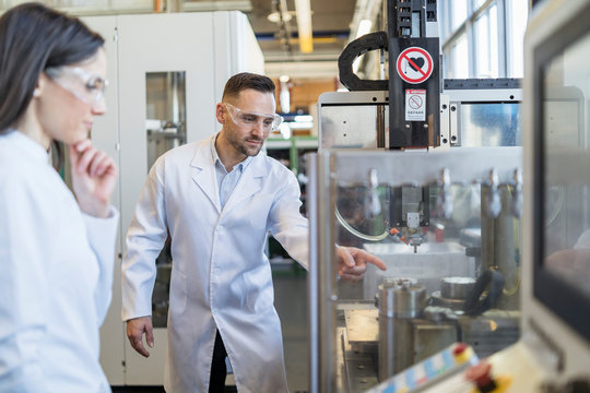 Colleagues Wearing Lab Coats And Safety Goggles Looking At Machine In Modern Factory