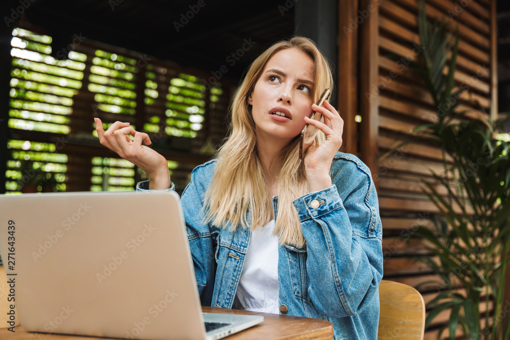 Wall mural Confused sad young woman posing outdoors in cafe using laptop computer and talking by mobile phone.