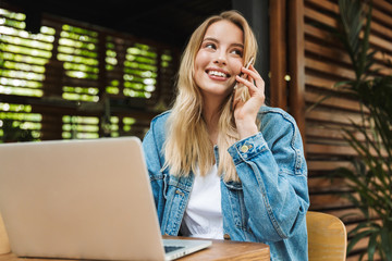Excited happy young woman posing outdoors in cafe using laptop computer and talking by mobile phone.