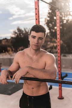 Young shirtless guy leaning on bar and looking at camera while working out on sports ground on sunny day on city street