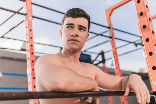 Young shirtless guy leaning on bar and looking at camera while working out on sports ground on sunny day on city street