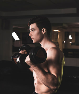 Strong young guy covered with electric discharges looking at camera while lifting heavy dumbbells in gym