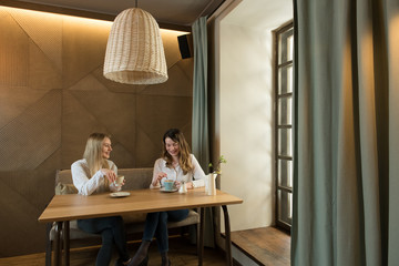 Two stylish female friends with cups of fresh coffee smiling and talking while sitting at table in cozy restaurant