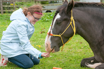 Young girl feeding carrot treats to a pony lying down on the grass.
